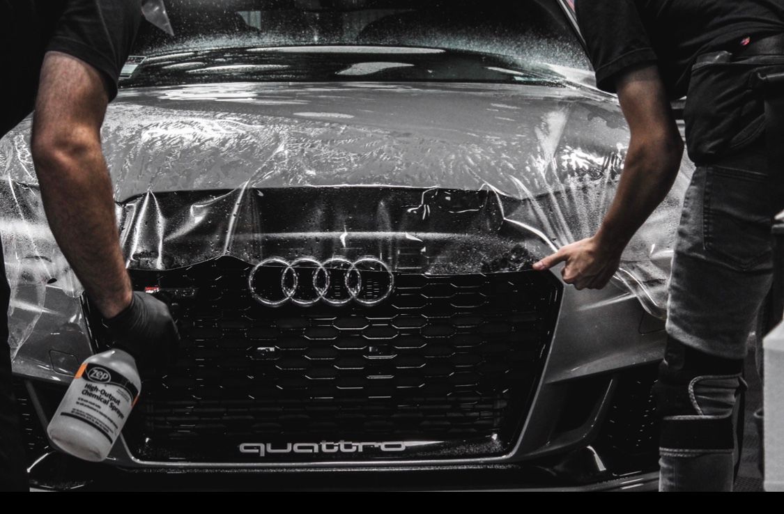 two workers pulling film over an Audi hood, spraying it with water for application